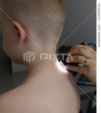 A pediatric dermatologist examining a young boys skin with a dermatoscope in a medical clinic. Skin check up for moles and birthmarks. A pediatric dermatologist examining a young boys skin with a dermatoscope in a medical clinic. Skin check up for moles and birthmarks. 129090628