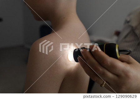 A pediatric dermatologist examining a young boys skin with a dermatoscope in a medical clinic. Skin check up for moles and birthmarks. A pediatric dermatologist examining a young boys skin with a dermatoscope in a medical clinic. Skin check up for moles and birthmarks. 129090629