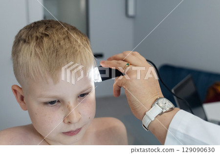 A pediatric dermatologist examining a young boys skin with a dermatoscope in a medical clinic. Skin check up for moles and birthmarks. A pediatric dermatologist examining a young boys skin with a dermatoscope in a medical clinic. Skin check up for moles and birthmarks. 129090630