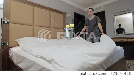 Hotel maid arranging white sheets, smoothing bedding while preparing room for incoming guests. Housekeeping service in hotel room. 129091498