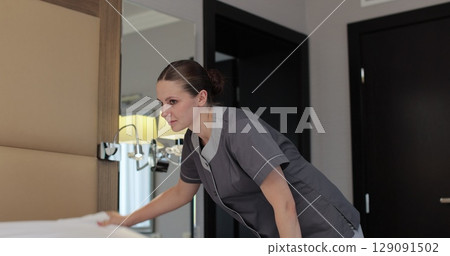 Young maid in professional uniform makes bed in hotel room adjusting white blanket. Cleaning service in hotel rooms, hotel business. 129091502