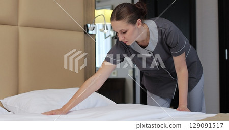 Female housekeeper in uniform making bed while preparing hotel room for guests. Hotel cleaning concept. Female housekeeper in uniform making bed while preparing hotel room for guests. Hotel cleaning concept. 129091517