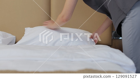 Close-up of young maid in uniform, fluffing pillows and punching them into shape. Hotel cleaning concept. 129091527