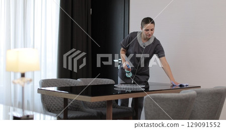 Housekeeper in uniform dusting table in modern hotel room while performing daily cleaning service. Cleaning service in hotel rooms, hotel business. 129091552