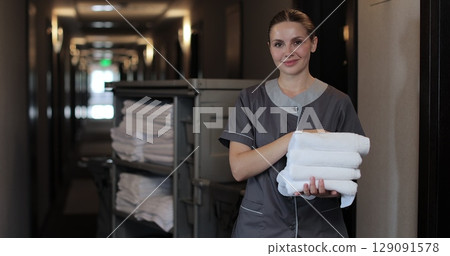 Portrait of young maid with trolley in hotel hallway, cleaning service, staff. Maid with clean white towels in hands. Daily cleaning. Cleaning service in hotel rooms, hotel business. Portrait of young maid with trolley in hotel hallway, cleaning service, staff. Maid with clean white towels in hands. Daily cleaning. Cleaning service in hotel rooms, hotel business. 129091578