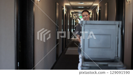 Maid rolling trolley in corridor of modern hotel, cleaning service in hospitality. Daily cleaning. Cleaning service in hotel rooms, hotel business. Maid rolling trolley in corridor of modern hotel, cleaning service in hospitality. Daily cleaning. Cleaning service in hotel rooms, hotel business. 129091590