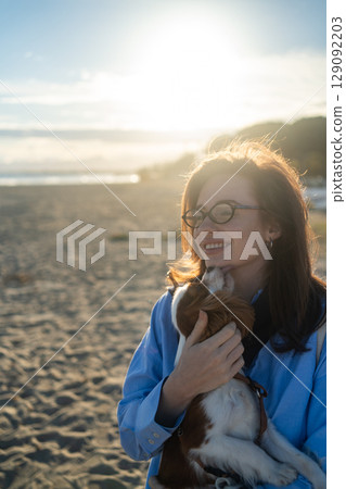 A young woman hugs her Cavalier King Charles Spaniel on the beach at sunset. Warm light, waves, and smiles capture a peaceful, joyful moment of connection and relaxation. 129092203