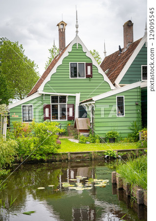 Dutch typical landscape. Traditional old dutch windmill with old houses against blue cloudy sky in the Zaanse Schans village, Netherlands. Famous tourism place. Sheep grazing on green grass. 129095545