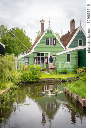 Dutch typical landscape. Traditional old dutch windmill with old houses against blue cloudy sky in the Zaanse Schans village, Netherlands. Famous tourism place. Sheep grazing on green grass. Dutch typical landscape. Traditional old dutch windmill with old houses against blue cloudy sky in the Zaanse Schans village, Netherlands. Famous tourism place. Sheep grazing on green grass. 129095546
