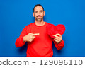 Bearded Hispanic man in his 40s wearing a red sweater holding a red heart as a symbol of romance and health, looking surprised and pointing at himself with his finger. Isolated on a blue background. 129096110