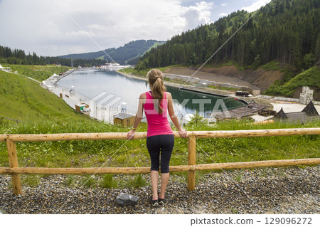 Back view of a young sportive woman in fitness clothes standing outdoors near big lake in mountains enjoying nature view. Back view of a young sportive woman in fitness clothes standing outdoors near big lake in mountains enjoying nature view. 129096272