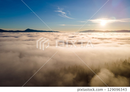 Aerial view of vibrant sunset over white dense clouds with dark mountain spruce forest trees. Aerial view of vibrant sunset over white dense clouds with dark mountain spruce forest trees. 129096343