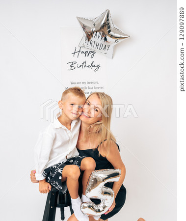 Happy mom hugging her 5-year-old son at birthday celebration with festive balloons and decorations in bright indoor setting 129097889