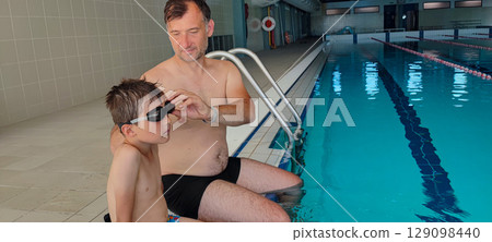 A father helps his son with swimming goggles in a large indoor pool. Concept: strengthening family bonds through training, joint sports, fatherhood. 129098440