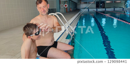 A father helps his son with swimming goggles in a large indoor pool. Concept: strengthening family bonds through training, joint sports, fatherhood. A father helps his son with swimming goggles in a large indoor pool. Concept: strengthening family bonds through training, joint sports, fatherhood. 129098441