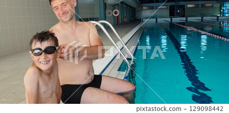A father helps his son with swimming goggles in a large indoor pool. Concept: strengthening family bonds through training, joint sports, fatherhood. 129098442