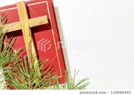 Wood cross laying on a closed red Christian bible with green fir branches on a white textile background with copy space. Christmas holidays. Christian religion background. Flat lay 129098449