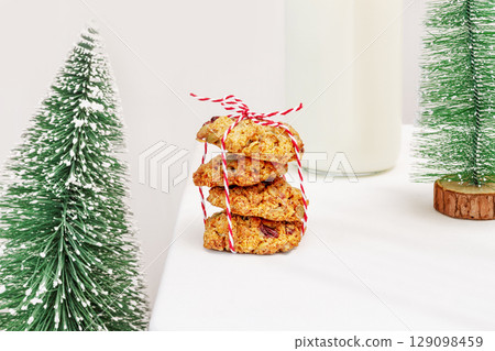 Christmas and New Year still life. Winter holidays food composition with natural handmade cookies with seeds and cranberries, bottle of milk and small new year trees on a white table Christmas and New Year still life. Winter holidays food composition with natural handmade cookies with seeds and cranberries, bottle of milk and small new year trees on a white table 129098459