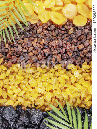 Dry fruits assortment with palm leaves. Rows of scattered dried apricots, brown raisins, golden raisins and prunes. Healthy snack - mix of organic dry fruits. Vegan and vegetarian products. Flat lay Dry fruits assortment with palm leaves. Rows of scattered dried apricots, brown raisins, golden raisins and prunes. Healthy snack - mix of organic dry fruits. Vegan and vegetarian products. Flat lay 129098593