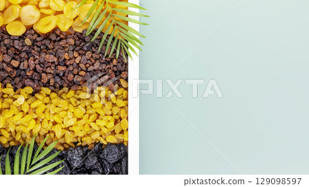 Dry fruits assortment. Rows of scattered dried apricots, brown raisins, golden raisins and prunes on white wooden tray on blue background with copy space. Healthy snack - mix of organic dry fruits Dry fruits assortment. Rows of scattered dried apricots, brown raisins, golden raisins and prunes on white wooden tray on blue background with copy space. Healthy snack - mix of organic dry fruits 129098597