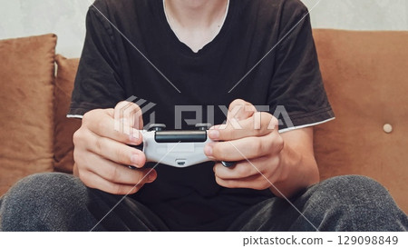 Joystick in hand: a person spends time playing games, a relaxed position. Gaming focused relaxation. silhouette of a man - a teenage boy playing a video game. Joystick in hand: a person spends time playing games, a relaxed position. Gaming focused relaxation. silhouette of a man - a teenage boy playing a video game. 129098849