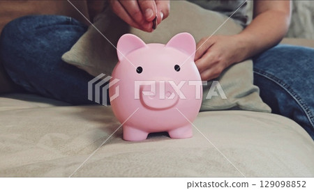 Woman saving cash in bank. Close up of woman placing coins into piggy bank. Concept of financial planning. Piggy bank symbolizes saving cash. Woman managing finances. Budgeting and financial security. 129098852