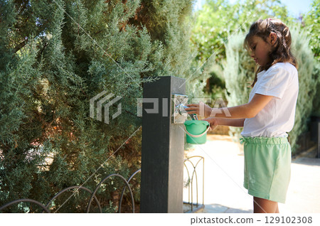 Young Girl Filling a Container From a Garden Water Tap Surrounded by Lush Trees 129102308