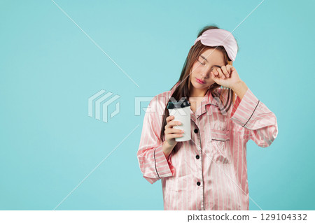 Young woman in pajamas with eye mask holding coffee cup after waking up, she needs more sleep, walking to get some coffee, still needs some sleep, headshots isolated studio blue background Young woman in pajamas with eye mask holding coffee cup after waking up, she needs more sleep, walking to get some coffee, still needs some sleep, headshots isolated studio blue background 129104332