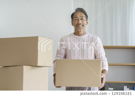 Senior man holding a cardboard box and smiling while looking at the camera. Image of moving and decluttering. 129106181