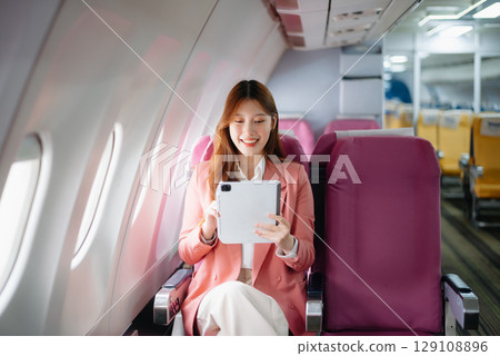 Portrait of a successful Asian businesswoman or entrepreneur in a formal suit on an airplane sitting in business class using a phone, computer laptop. Travel in style, work 129108896