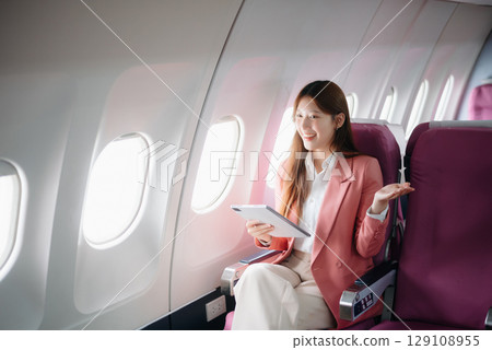 Portrait of a successful Asian businesswoman or entrepreneur in a formal suit on an airplane sitting in business class using a phone, computer laptop. Travel in style, work 129108955