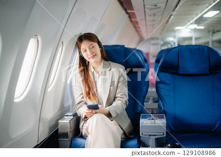 Portrait of a successful Asian businesswoman or entrepreneur in a formal suit on an airplane sitting in business class using a phone, computer laptop. Travel in style, work 129108986