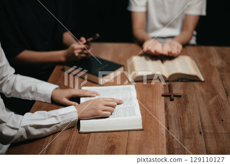 Group of Christians sit together and pray around a wooden table with blurred open Bible pages in their homeroom. 129110127