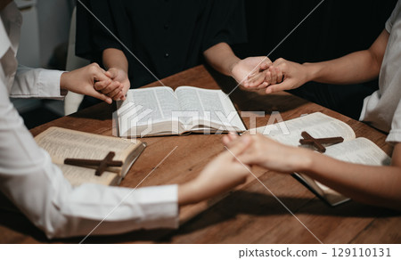Group of Christians sit together and pray around a wooden table with blurred open Bible pages in their homeroom. 129110131