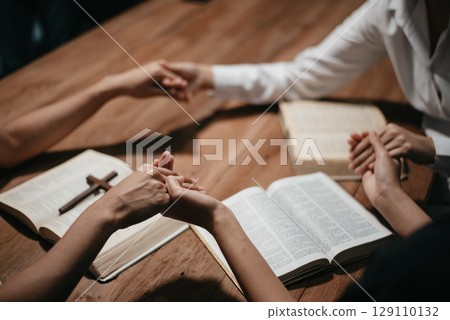Group of Christians sit together and pray around a wooden table with blurred open Bible pages in their homeroom. 129110132