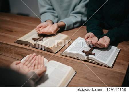 Group of Christians sit together and pray around a wooden table with blurred open Bible pages in their homeroom. Group of Christians sit together and pray around a wooden table with blurred open Bible pages in their homeroom. 129110138