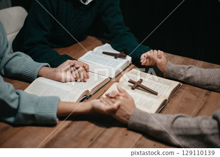 Group of Christians sit together and pray around a wooden table with blurred open Bible pages in their homeroom. 129110139