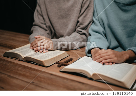 Group of Christians sit together and pray around a wooden table with blurred open Bible pages in their homeroom. 129110140