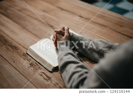 Group of Christians sit together and pray around a wooden table with blurred open Bible pages in their homeroom. 129110144