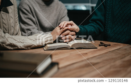 Group of Christians sit together and pray around a wooden table with blurred open Bible pages in their homeroom. 129110146