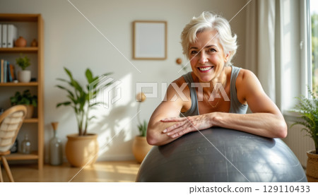 Smiling senior woman with short white hair leaning on exercise ball in bright room with plants and wooden furniture, feeling happy and healthy 129110433