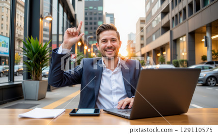 Young businessman smiling and raising hand while working on laptop outdoors in city street at sunset 129110575