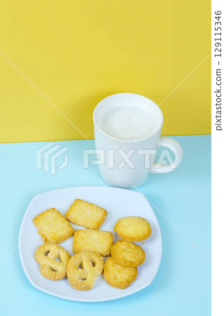 A plate of various shortbread cookies and a white mug of milk sit on a blue surface against a yellow wall. A plate of various shortbread cookies and a white mug of milk sit on a blue surface against a yellow wall. 129115346