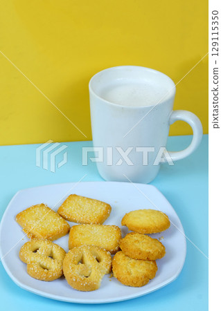 A plate of various shortbread cookies and a white mug of milk sit on a blue surface against a yellow wall. A plate of various shortbread cookies and a white mug of milk sit on a blue surface against a yellow wall. 129115350