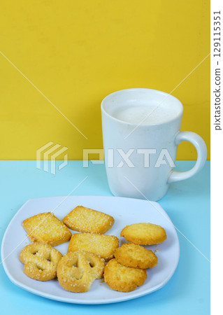 A plate of various shortbread cookies and a white mug of milk sit on a blue surface against a yellow wall. 129115351