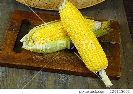 Two fresh, yellow corns, one partially husked, rest on a rustic wooden cutting board with a woven basket in the background. 129115661