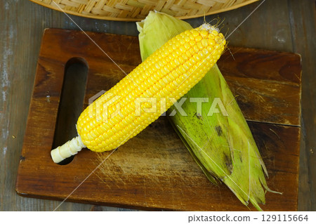 Two fresh, yellow corns, one partially husked, rest on a rustic wooden cutting board with a woven basket in the background. Two fresh, yellow corns, one partially husked, rest on a rustic wooden cutting board with a woven basket in the background. 129115664