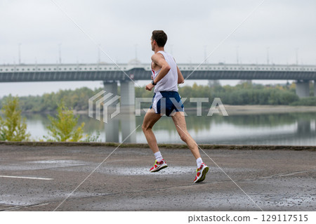 runner wearing sleeveless shirt and shorts, captured on cloudy day with large bridge and river 129117515