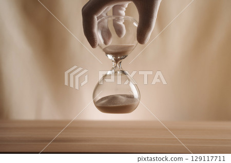 A glove-covered hand holding an hourglass on a wooden surface, symbolizing the flow of time, creativity, and patience in a contemporary minimalist style. 129117711