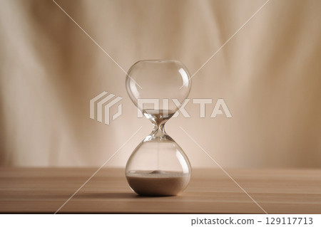 A simple hourglass on a wooden table with a beige backdrop, symbolizing time flow and minimalistic living. Running out of time, uncluttered presentation for stylish concepts. 129117713